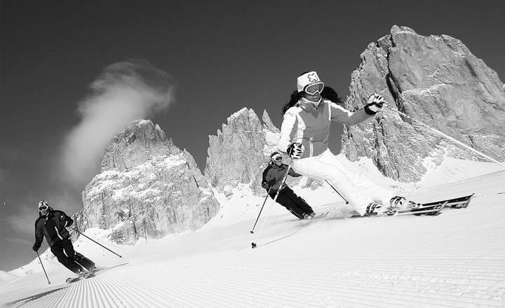Skiløbere på hvid piste med bjerge i baggrunden i Canazei på skiferie